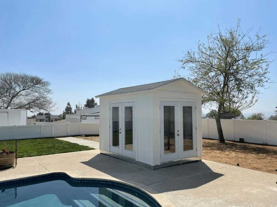 Small white shed with glass doors beside a pool, surrounded by a concrete patio, a grassy area, and trees under a clear blue sky.