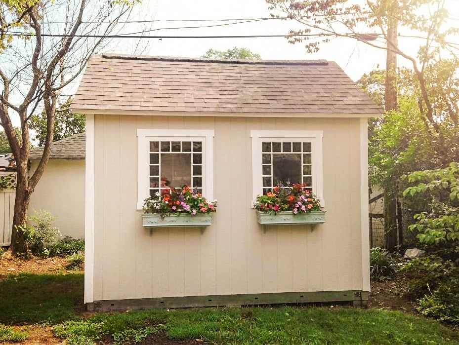 A small beige shed with a gabled roof, and two windows adorned with flower boxes filled with pink and white flowers.