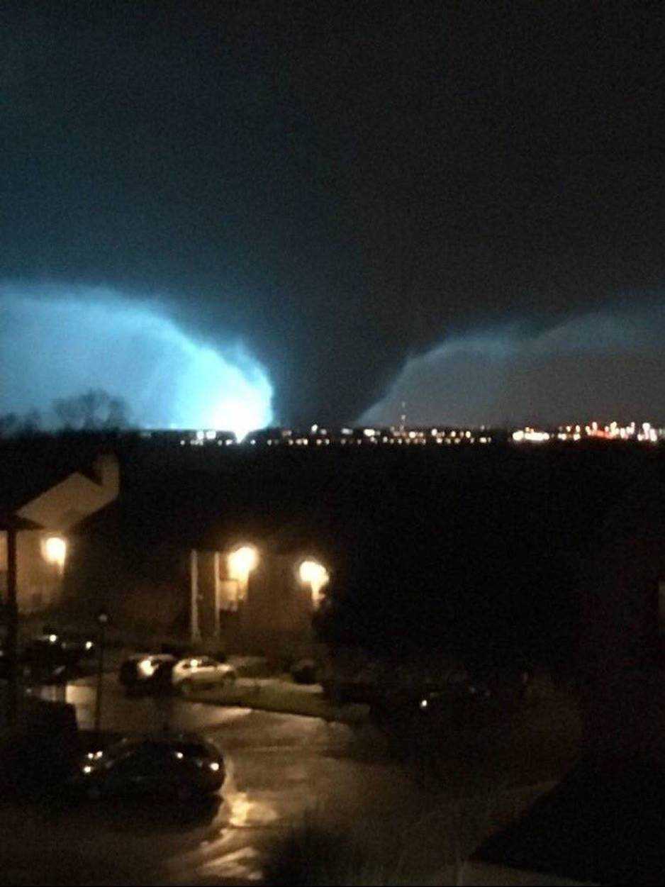 A photo taken during the storm A large tornado illuminated by lightning at night, moving towards a city with visible streetlights and buildings in the foreground