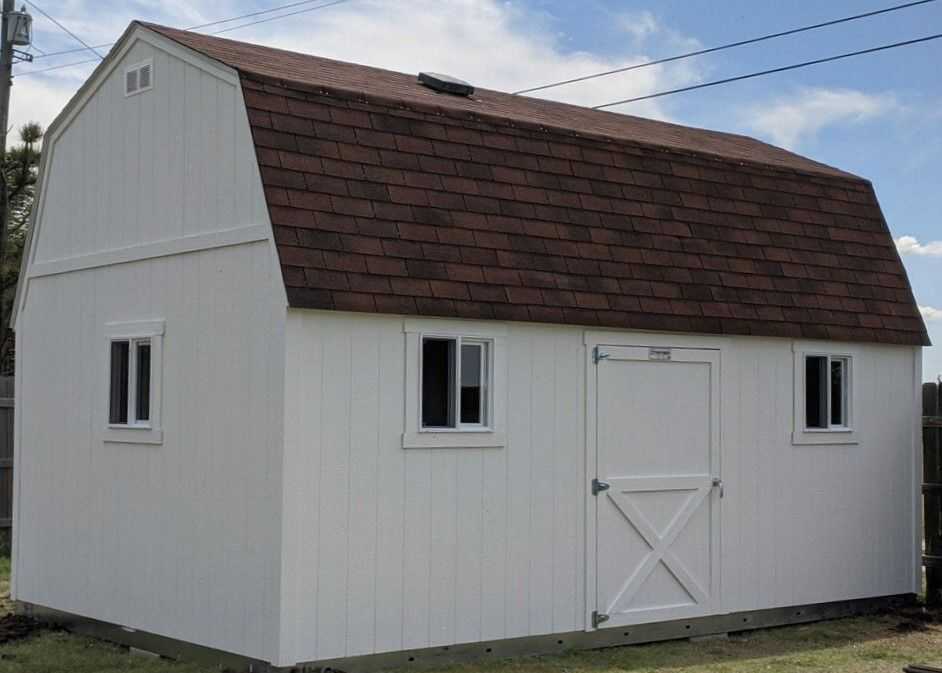 A white TB700 barn with a brown gambrel roof, two small windows, and a single door, set in a grassy yard under a blue sky.
