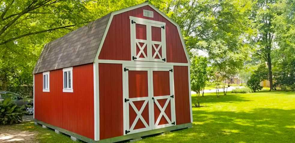 A red TB-700 barn-style shed with white trim and double doors stands on a lush green lawn surrounded by trees.